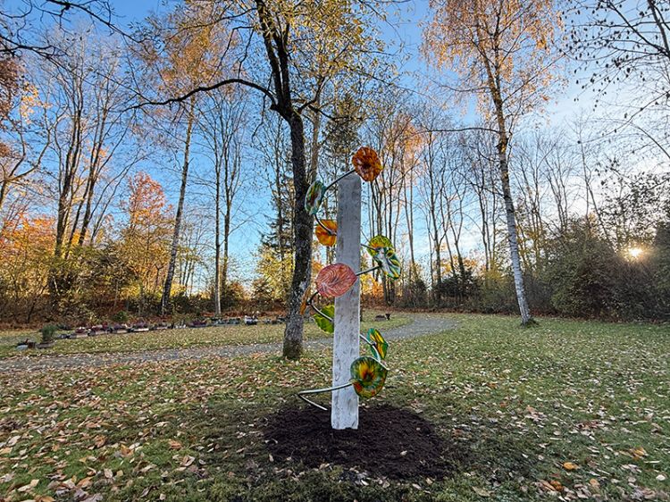 Weiße, schlanke Stele mit bunten, Glas-Blättern in einer herbstlichen Parklandschaft mit Bäumen und Laub auf dem Boden.