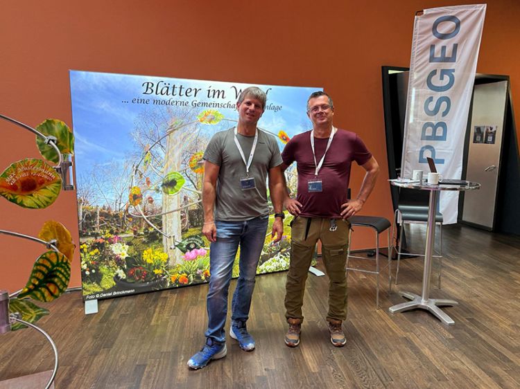 Zwei Männer mit Namensschildern stehen vor einem Banner mit der Aufschrift 'Blätter im Wind ... eine moderne Gemeinschaftsgrabanlage'. Rechts ein Stehtisch mit einem Banner 'PBSGEO'.