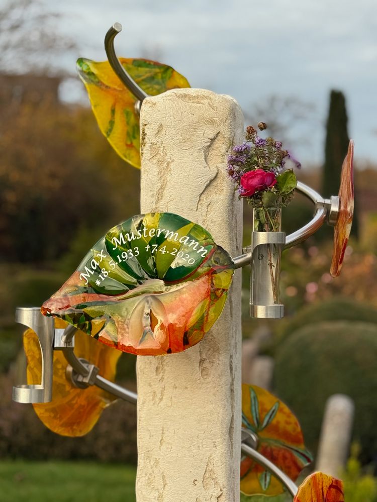 Grabstein mit bunten Glasblättern und der Inschrift 'Max Mustermann 18.8.1933 17.4.2020', daneben ein Metallvasenhalter mit frischen Blumen.